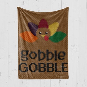 Puede incluir: Manta de crochet marrón con un diseño de pavo pixelado y el texto "gobble GOBBLE" en negro. La manta está colgada en una pared de madera blanca.