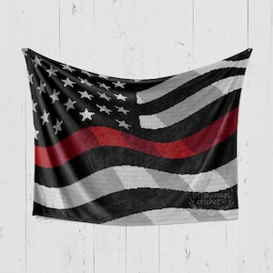 May include: A crocheted American flag blanket with a red stripe across the middle, representing the thin red line. The blanket is draped over a white wooden surface. The text "littlecraigs crochet" is visible in the bottom right corner.