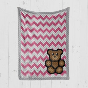 Puede incluir: Una manta de bebé de crochet en chevron rosa y gris con una aplicación de oso marrón en el centro.