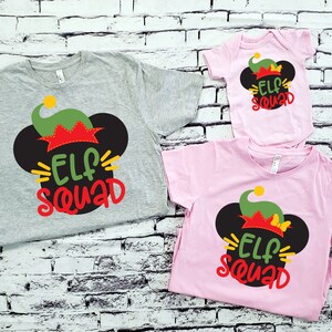 May include: Three shirts with a Christmas elf design. The shirts are grey, pink, and white. The design features a black Mickey Mouse ear with a green elf hat and the text "Elf Squad".