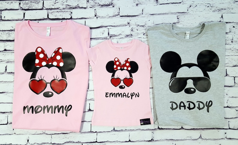 May include: Three pink and grey t-shirts with a Mickey Mouse and Minnie Mouse design. The pink shirts have a Minnie Mouse with red heart sunglasses and a bow. The grey shirt has a Mickey Mouse with black sunglasses. The shirts say "Mommy", "Emmalyn", and "Daddy".