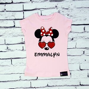 May include: Pink t-shirt with a black and red Minnie Mouse graphic wearing heart-shaped sunglasses. The shirt has the name "EMMALYN" printed below the graphic.