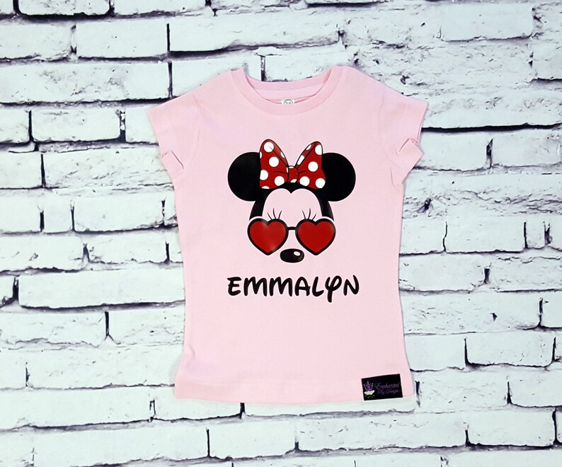 May include: Pink t-shirt with a black and red Minnie Mouse graphic wearing heart-shaped sunglasses. The shirt has the name "EMMALYN" printed below the graphic.