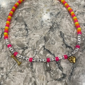 May include: A pink and yellow beaded necklace with the words "DO IT" and "WHACK" on it. The necklace has a gold bone charm and a silver paw print charm.