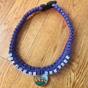 May include: A purple paracord necklace with white beads that spell "I AM BB SS IT BB TH" and a round charm with a nature scene.
