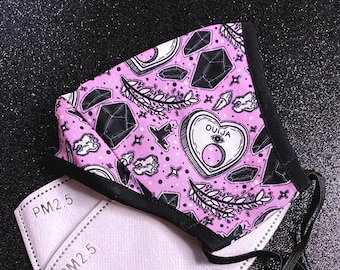 May include: A pink face mask with a black border and a black and white print of a Ouija board, crystals, and other occult symbols. Two white PM2.5 filters are included.