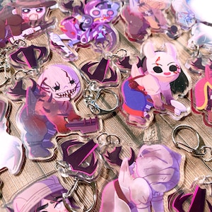 Puede incluir: Una colección de llaveros acrílicos transparentes con varios personajes de dibujos animados en tonos morados, rosas y marrones. Cada llavero tiene un llavero plateado adjunto. Los personajes tienen una variedad de expresiones y poses.