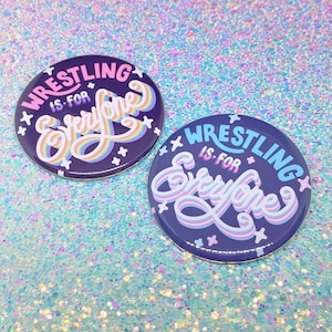 May include: Two circular, dark purple badges with the text "Wrestling is for Everyone" in a colourful, whimsical font. The badges have a glittery background and are likely pins.