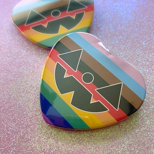 May include: Two heart-shaped rainbow pride pins with a black and white graphic of a smiling face with triangles for eyes and a circle for a nose.
