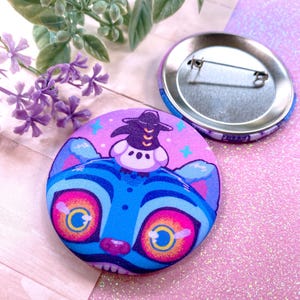 May include: A round button with a cartoon cat design. The cat is blue with large pink and yellow eyes, and a witch hat. The background is purple with stars. A second button is visible.