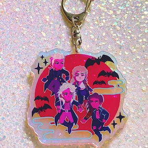 Puede incluir: Un llavero hologr&aacute;fico con cinco personajes de dibujos animados con cabello negro y piel rosa. Los personajes est&aacute;n de pie frente a una luna roja con murci&eacute;lagos volando a su alrededor. El llavero est&aacute; rodeado por un borde blanco con un efecto brillante.
