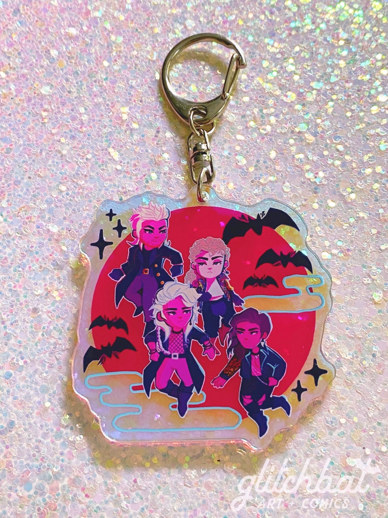 Puede incluir: Un llavero hologr&aacute;fico con cinco personajes de dibujos animados con cabello negro y piel rosa. Los personajes est&aacute;n de pie frente a una luna roja con murci&eacute;lagos volando a su alrededor. El llavero est&aacute; rodeado por un borde blanco con un efecto brillante.