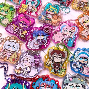 May include: A collection of colorful anime-style character keychains. Each keychain features a unique character design with vibrant colors, glitter accents, and a beaded chain for attachment. The characters are depicted in a cute, chibi style.