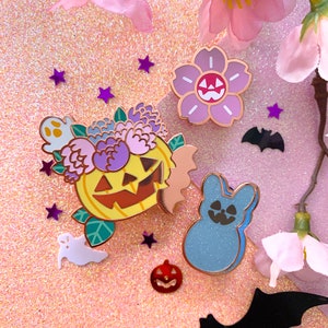 May include: A set of five enamel pins featuring a pumpkin with a flower crown, a blue bunny, a pink flower with a jack-o-lantern face, a ghost, and a bat. The pins are on a pink glitter background.