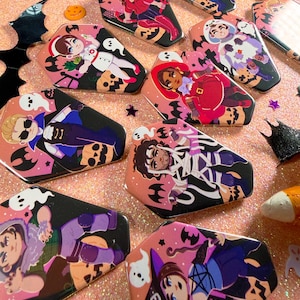 May include: A collection of coffin-shaped pins featuring various illustrated characters in Halloween costumes, including a mummy, a vampire, a nurse, and a witch, all accented with small ghost and pumpkin motifs on a glittery pink surface.