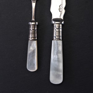 May include: A silver butter knife and fork with mother-of-pearl handles. The knife has a decorative floral design on the blade.