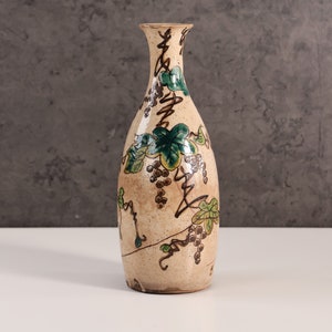 May include: A ceramic vase with a cream-colored background and a floral design featuring green leaves and brown grapes.