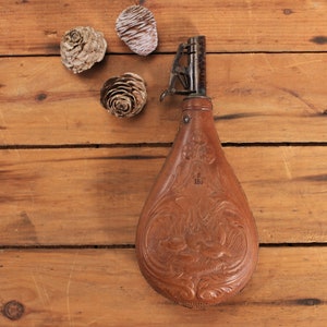 May include: A brown leather powder flask with intricate embossed designs and a metal spout. The flask is on a wooden surface with pine cones.