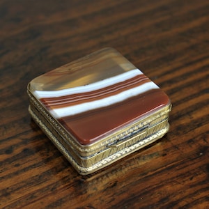 May include: A small, square, gold-toned box with a hinged lid. The lid is made of agate with brown, white, and red stripes.