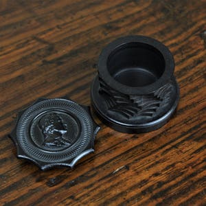 May include: A black wooden box with a carved design and a separate lid with a raised profile. The lid features a carved image of a person's head.