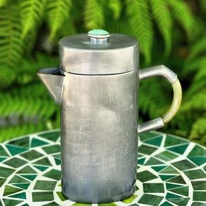 Könnte beinhalten: Eine silberne French Press Kaffeemaschine mit grünem Deckel und hellbraunem Griff steht auf einem grün-weißen Mosaiktisch.