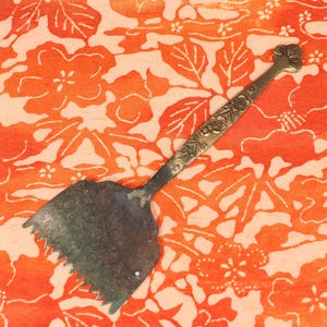 May include: A silver-colored metal cake server with a decorative floral design and a serrated edge. The server is lying on a red and white floral fabric.