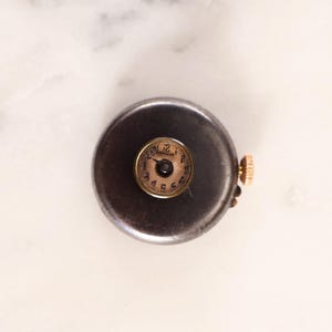May include: A small, antique pocket watch with a dark, circular casing. The watch face is a light brown color with black numerals and hands. A gold-colored crown is visible on the side.