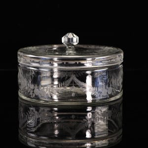 Peut inclure: Un bocal en verre transparent avec un couvercle, orné de motifs floraux et de feuilles gravés. Le bocal est de forme ronde et possède un bouton à facettes sur le dessus. Le bocal est posé sur une surface réfléchissante.