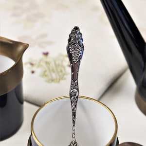 Puede incluir: Una cucharadita de plata con un diseño de vid se encuentra dentro de una taza de té azul y dorada con un asa de latón.