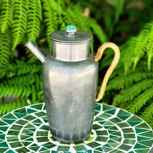 Könnte beinhalten: Ein silberner Cocktail-Shaker aus Zinn mit einem grünen Stein oben und einem geflochtenen Griff. Der Shaker steht auf einem grünen Mosaiktisch.