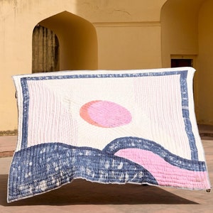 May include: A rectangular quilt with a landscape design featuring a pink sun, blue waves, and white background is held up by two people. The quilt is displayed in front of a yellow building with arched windows and a large, curved structure. The people are wearing traditional Indian clothing.