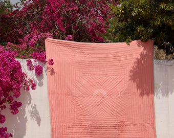 May include: A coral-colored, quilted blanket hangs against a white wall, with a geometric design in the center. Lush pink bougainvillea and green trees frame the scene under a clear blue sky. The blanket is square-shaped.