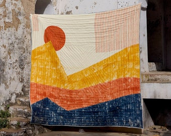 May include: A colorful quilt with a geometric design featuring a red sun, yellow and orange mountains, and blue water. The quilt is hanging on a white wall with a stone archway in the background.
