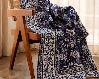 Peut inclure: Une chaise en bois avec un dossier et une assise en cannage, recouverte d'un textile rectangulaire à motif floral bleu foncé et blanc. Le tissu présente une large bordure décorative et de petits pompons blanc cassé aux coins inférieurs.
