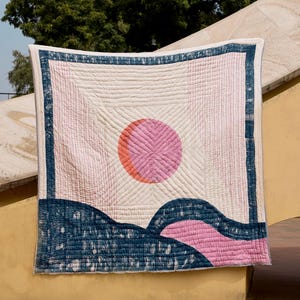 May include: A quilted textile with a geometric design. The quilt features a pink and orange sun, blue and pink landscape elements, and a blue border on a cream background. The quilt is hanging outdoors against a blue sky and trees.
