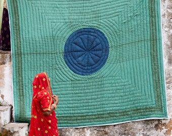 May include: A large, teal and blue quilt with a circular design is held up by three people. The quilt has a geometric pattern and is bordered by a darker teal. The people are wearing traditional Indian clothing.