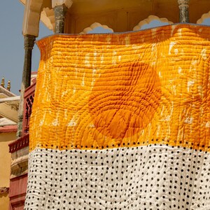 May include: A yellow and white patterned quilt hangs from a yellow building with a white trim. The quilt has a large yellow section with a white pattern and a white section with black polka dots.