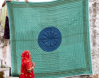 May include: Three women in traditional Indian clothing are holding up a large, green quilted blanket with a blue circle design in the center. The blanket is hanging on a white wall.