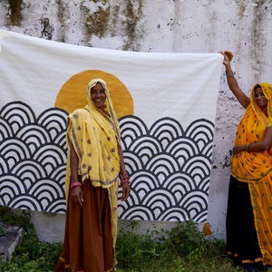 Peut inclure: Un textile rectangulaire blanc pr&eacute;sentant un grand motif de soleil jaune au-dessus de motifs de vagues en arc gris fonc&eacute; r&eacute;p&eacute;titifs. Le tissu pr&eacute;sente des coutures complexes sur toute sa surface, cr&eacute;ant une texture au design graphique &eacute;pur&eacute;.
