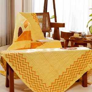 Puede incluir: Una colcha kantha amarilla y naranja con un patr&oacute;n en zigzag sobre un taburete de madera. La colcha est&aacute; doblada y drapeada sobre una silla en el fondo.