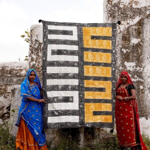 May include: Two people stand in front of a large, black and white patterned fabric with a yellow section. The fabric has a geometric design. The person on the left is wearing a blue and orange sari. The person on the right is wearing a red sari.