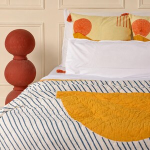 May include: A bed with white sheets and a striped comforter featuring a yellow half-circle design. Two yellow pillows with orange sunburst patterns and tassels are visible. A tall, red decorative object stands to the left.