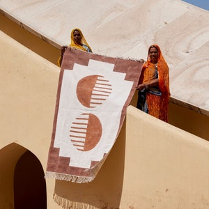 May include: Two women in colorful saris stand on a beige wall holding a large, rectangular rug with a brown and white geometric pattern. The rug has two large, half-circle shapes with a striped pattern.