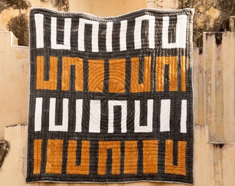 May include: A black and white geometric patterned quilt with orange squares. The quilt is hanging on a wall with a stone archway in the background.