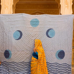 Può includere: Una coperta trapuntata azzurra con un motivo di fasi lunari blu scuro e linee ondulate sul bordo inferiore. Il copriletto presenta una texture impunturata su tutta la superficie ed è mostrato davanti a uno sfondo architettonico.