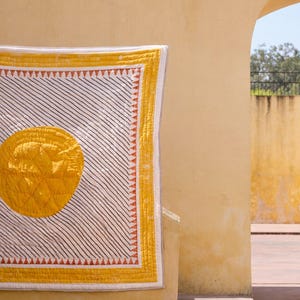 May include: A yellow and white quilt with a large yellow circle in the center. The quilt has black diagonal stripes and a yellow border with orange triangles. The quilt is hanging on a yellow wall.