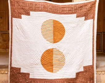 May include: A white and brown quilt with a geometric pattern of two large circles. The circles are divided in half, with one half being a solid brown colour and the other half having a striped pattern. The quilt is hanging on a wall with a yellow background.