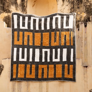 May include: A black and white geometric patterned quilt with orange squares. The quilt is hanging on a wall with a stone archway in the background.