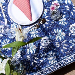 Puede incluir: Un mantel de mesa floral azul y blanco con una servilleta a cuadros rojos y blancos sobre un plato blanco. Hay lirios blancos en un jarr&oacute;n transparente sobre la mesa.