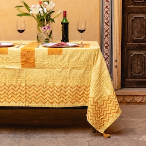 Puede incluir: Un mantel amarillo con un patrón geométrico en naranja, preparado para una cena romántica con dos copas de vino, una botella de vino y platos. El mantel tiene un borla en el extremo.